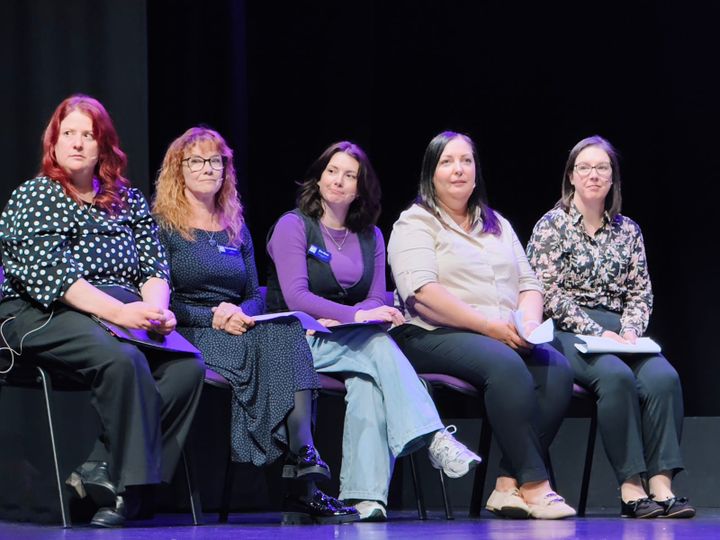 Five women seated on a stage, engaged and holding papers, wearing casual and formal outfits.