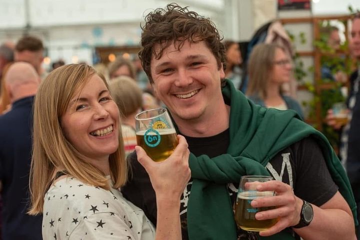 A man and woman stand close together indoors, smiling and holding glasses of beer with festival branding, surrounded by other attendees.