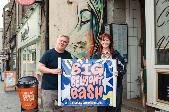 Two people holding a Big Belmont Bash banner outside a shop with mural art on the wall behind them.
