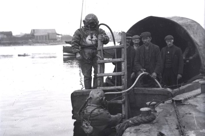 Black and white photo from around 1912 showing divers in heavy suits and helmets. One diver is in the water, another stands on the boat, with men behind.