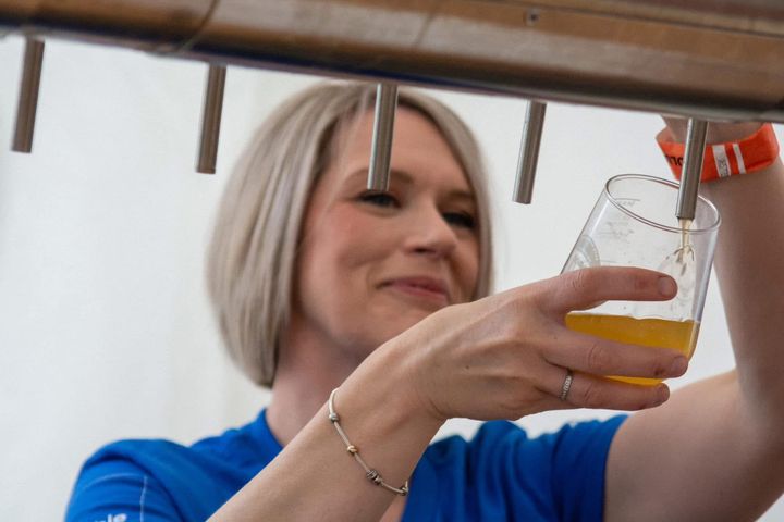 Woman in blue top pours beer from a tap into a glass at a festival, wearing a wristband.
