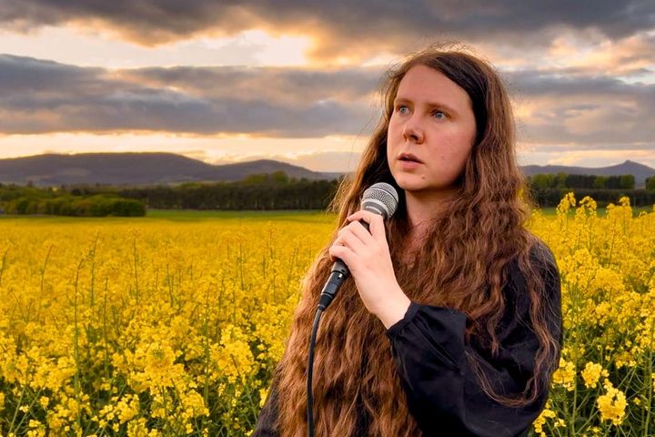 Lucy Beth holds a mic in a yellow field at sunset, performing in Doric for Ma Name Is Isabelle.