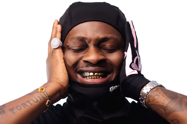 Close-up of PsychoYP in a black head covering, eyes closed, hands on head, showing gold teeth.