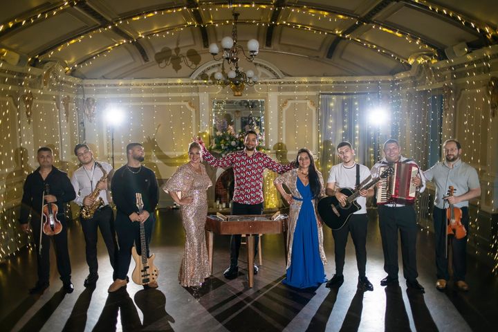 A group portrait of The Ando Glaso Collective, featuring nine musicians and singers dressed in formal attire. They stand beneath ornate chandeliers and strings of fairy lights.