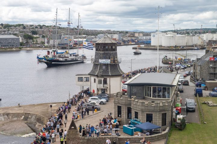 A tall ship decorated with flags sails past the historic Roundhouse in Aberdeen, while crowds line the pier and quayside below.