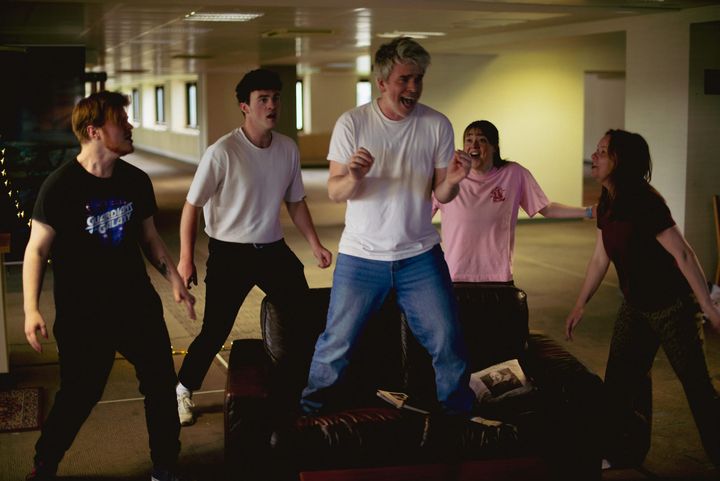 Five actors rehearse tick, tick… BOOM! with energy, one standing on a sofa mid-performance.