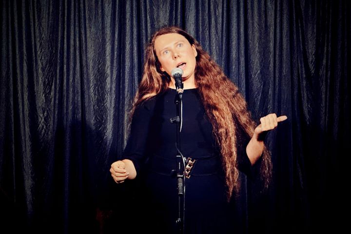 Lucy Beth performing on stage with long hair and black dress, speaking into a microphone.