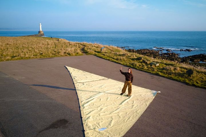 Laura Sherrifs standing on a large triangular sail by the coast, with sea and lighthouse in background.