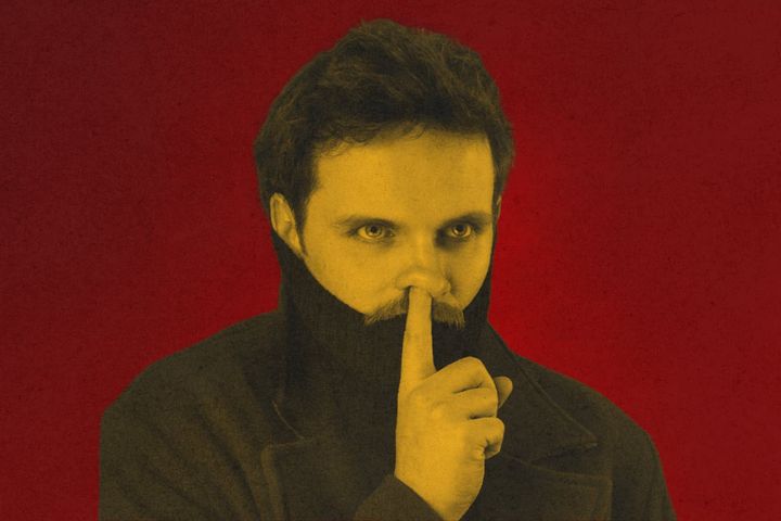 Comedian Vittorio Angelone stares ahead, finger raised to lips, set against a bold red background.
