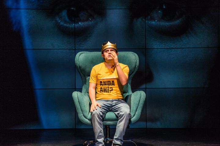 Actor wearing crown and yellow shirt sits on stage in front of large projected face.