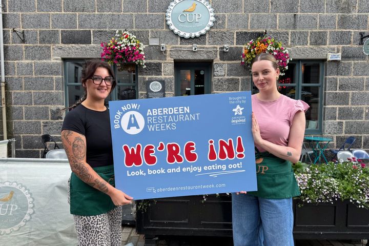 Two staff members from CUP in Aberdeen holding a sign reading “We’re In!” outside the café.