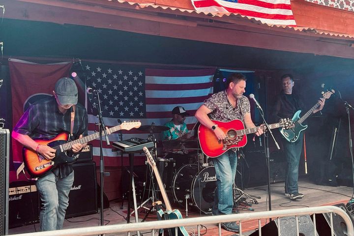 Four-piece band playing outdoors with American flags behind them.