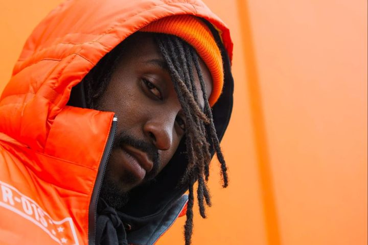 Close-up portrait of musician KAYO wearing an orange jacket and hat against an orange background.