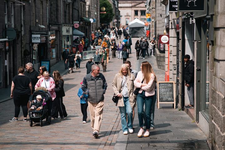 People stroll and chat along a sunlit Belmont Street during a busy afternoon event.
