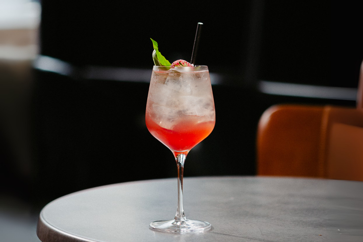 A tall glass of a red and clear cocktail with ice, basil and strawberry garnish, set on a table in soft light.