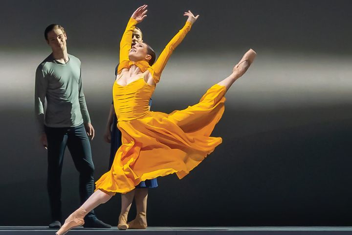 Dancer in flowing yellow dress performing a dramatic leap with arms raised and leg extended, whilst two figures in dark clothing watch from the background.