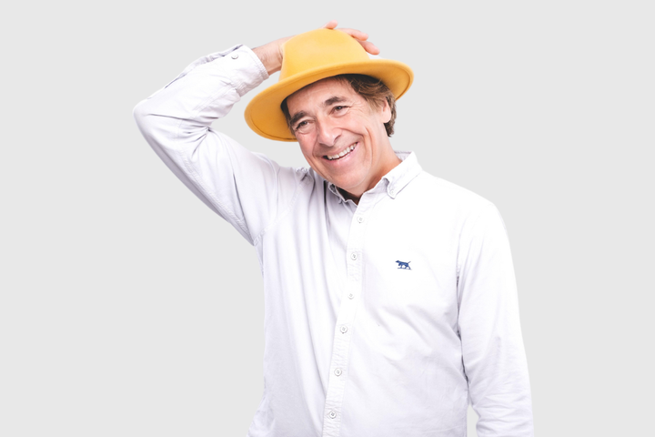 Mark Steel smiling in a white shirt and yellow hat, touching the top of his head against a pale grey background.