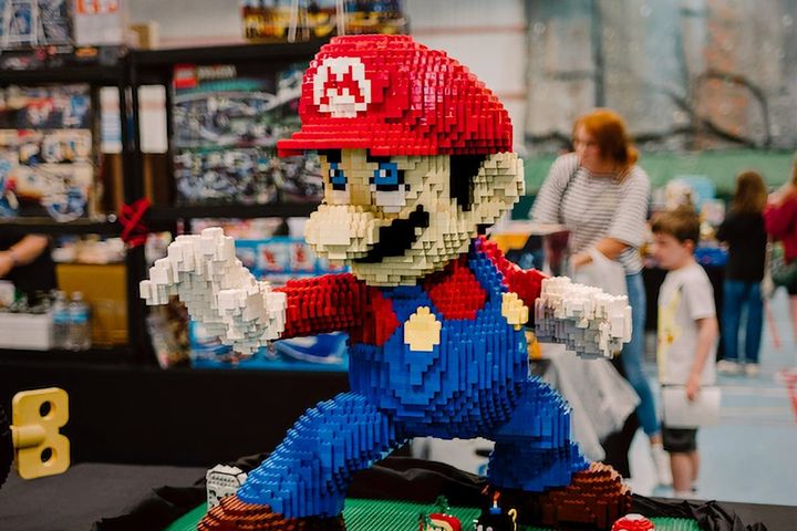 Large LEGO sculpture of Mario with red cap, moustache and blue dungarees displayed at indoor exhibition with shelves of LEGO products and visitors in the background.