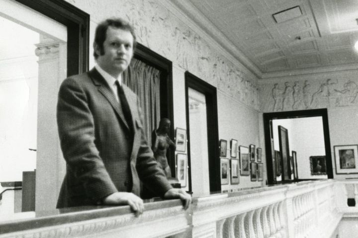 Black and white photo of Dr Ian McKenzie Smith standing by a balustrade inside Aberdeen Art Gallery, with framed artworks and a sculpture visible in the background.