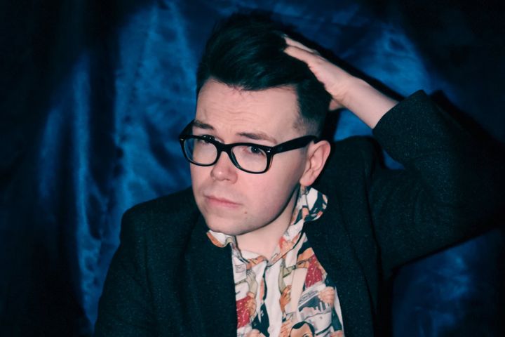 Christopher Macarthur-Boyd in glasses and blazer against blue backdrop, hand in hair