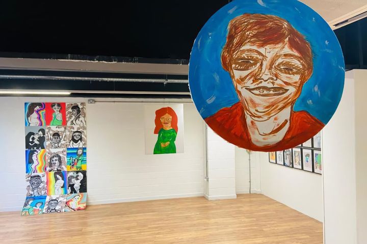 Gallery installation view showing large circular portrait painting with blue background and orange-red tones suspended from ceiling, grid of colourful portrait artworks on left wall.