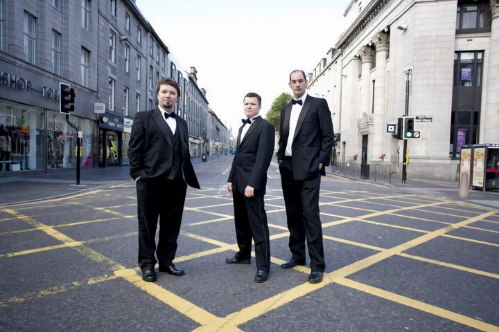 Ocean's 3 band members, three men in black tuxedos and bow ties, standing on yellow box junction markings in the middle of a quiet city street lined with grey stone buildings.