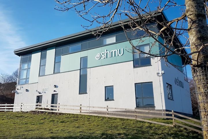 Modern two-storey shmu building with white walls and green upper section, photographed on a sunny day with bare tree branches in the foreground.