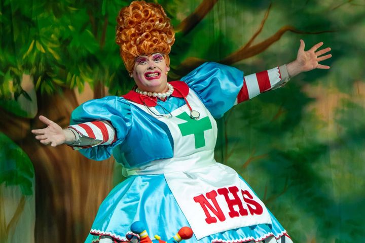 Pantomime dame in nurse costume with orange wig, arms wide.