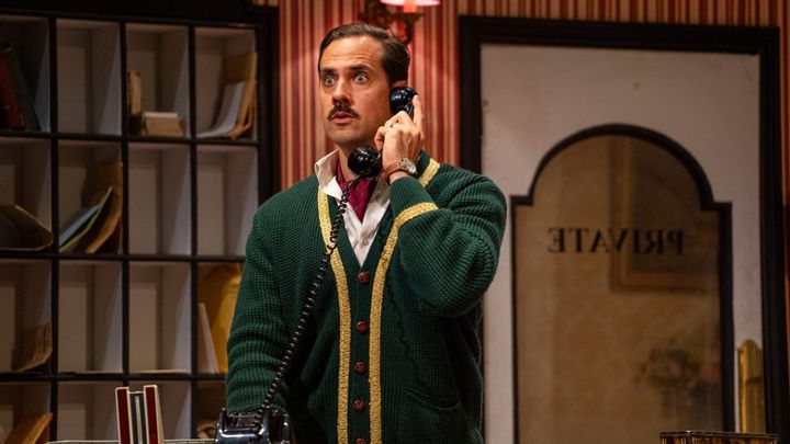 Basil on the phone at reception, eyes wide with alarm, wearing a green cardigan