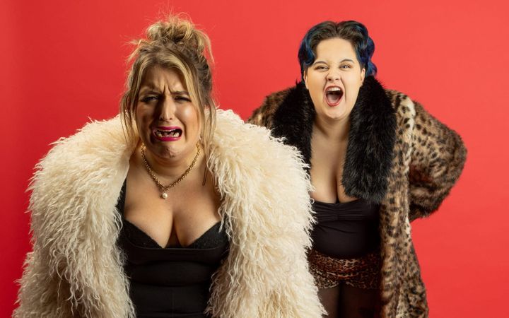 Two performers as the stepsisters in fur coats, pulling exaggerated faces against a red backdrop.