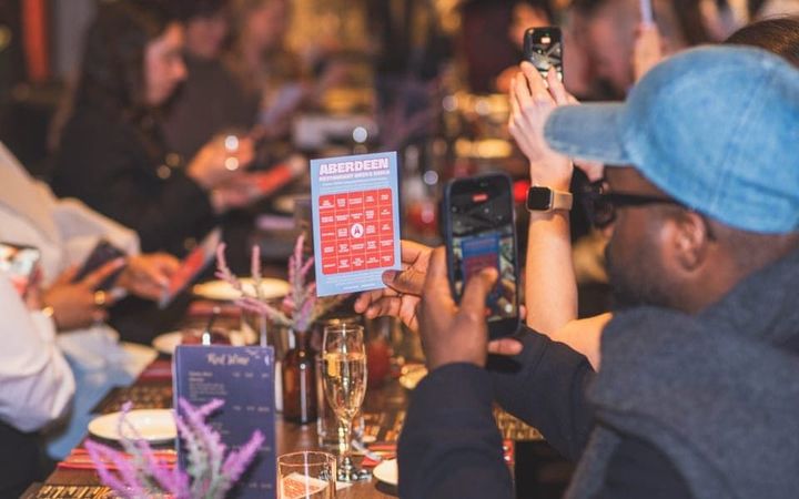 Diners at a restaurant table photographing the Aberdeen Restaurant Weeks bingo card