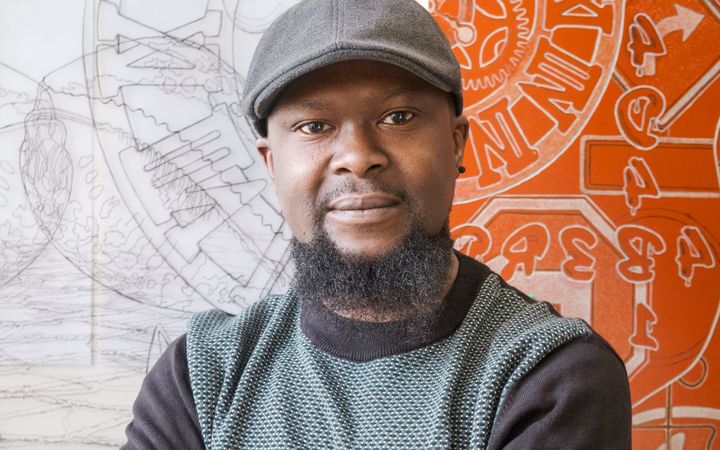 Artist Ade Adesina wearing a flat cap, standing in front of his detailed line drawings.