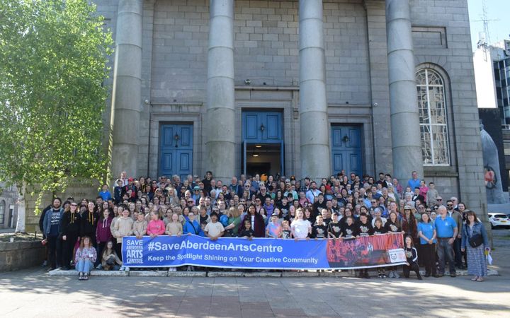 A large crowd on the steps of Aberdeen Arts Centre holding a Save Aberdeen Arts Centre banner.