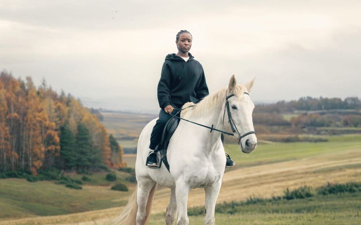 A young woman in a black hoodie rides a white horse across an open autumn landscape.