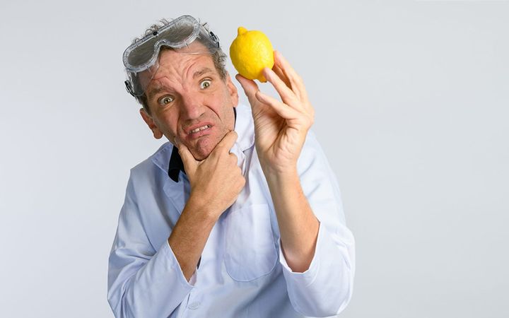 Comedian Henning Wehn in lab coat and goggles, holding a lemon with a puzzled expression.