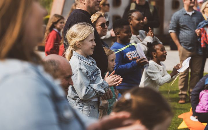 Children and families clapping and watching a performance outdoors on a sunny day