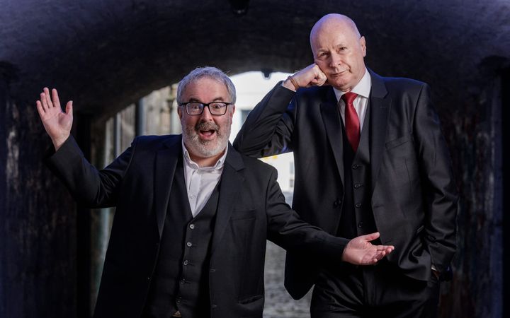 Two men in dark suits pose theatrically in a stone archway