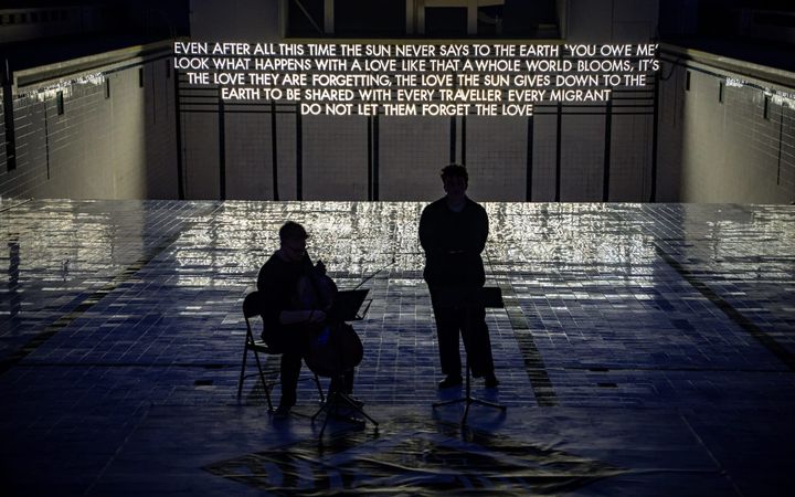 Two performers, one on cello, beneath Robert Montgomery’s lit poem at Bon Accord Baths