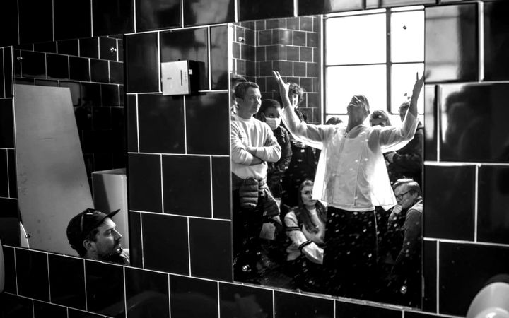 Alan Greig performing with arms raised, reflected in a row of bathroom mirrors.