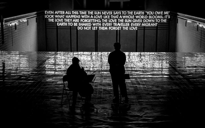 Two performers, one on cello, beneath Robert Montgomery’s lit poem at Bon Accord Baths