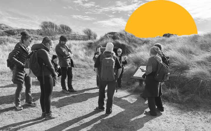 Group gathered on grassy dunes reading an information board, black and white with orange sun graphic.