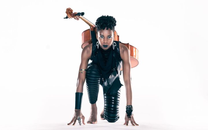 An athletic Ayanna Witter-Johnson on her knees like she was at the start of a race. She has her cello strapped to her back.