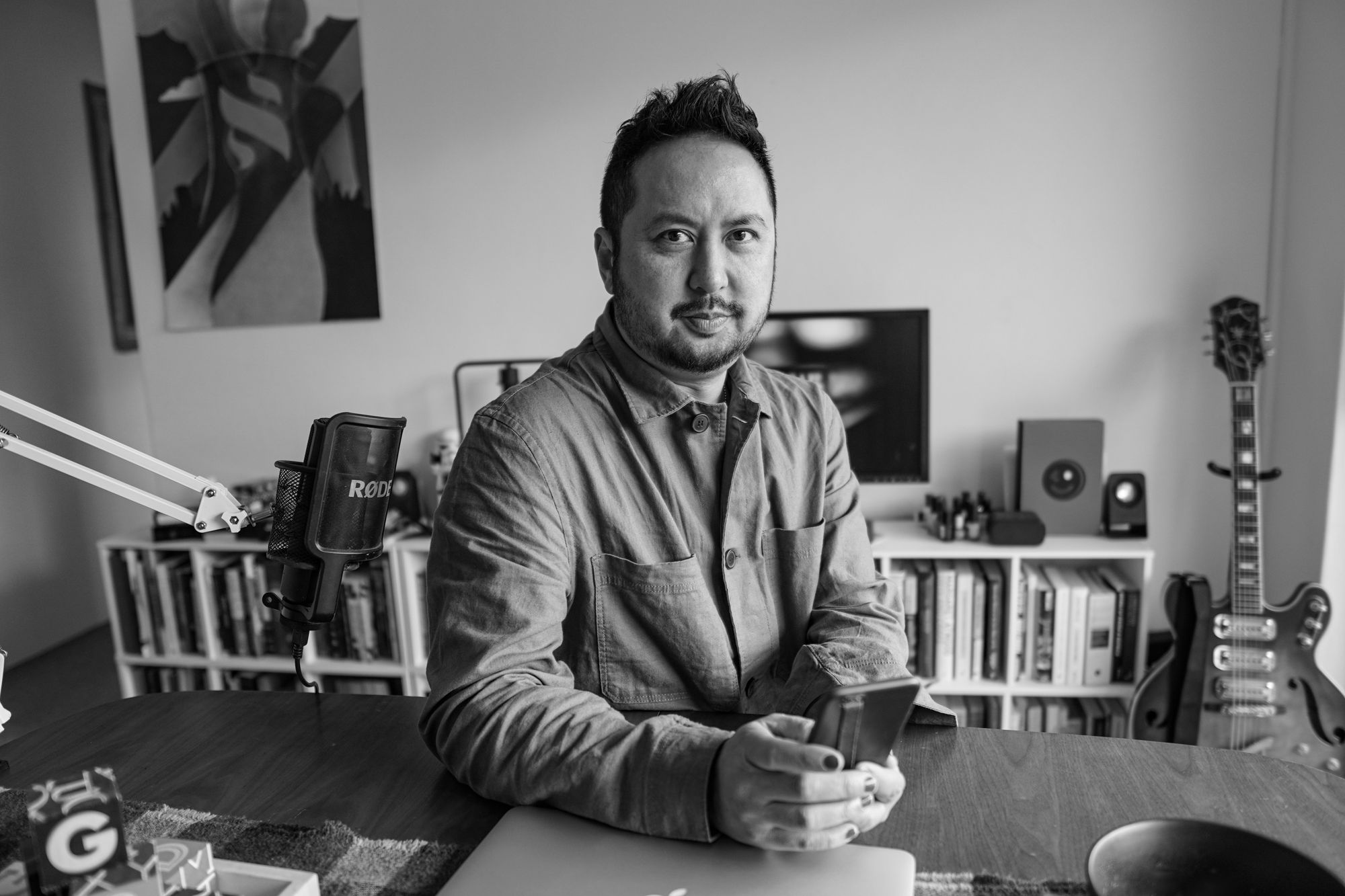 Portrait of Ben Hoh, sitting at his dining table next to a microphone, holding a mobile phone. In the background is a bookshelf and an electric guitar.