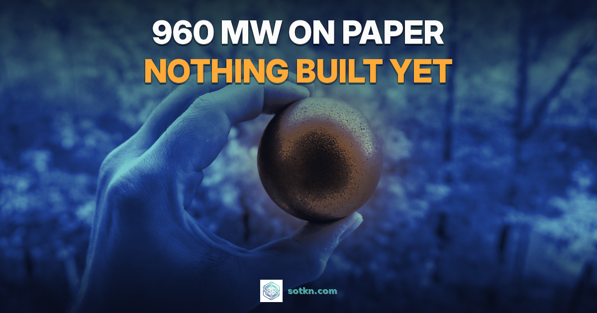 960 MW on paper, nothing built yet. A hand holding a dark graphite pebble of the kind used as reactor fuel, recoloured navy with an amber glow. Illustrative, not an X-energy part.