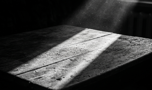 Crno-bela fotografija krupnog plana grubog, drvenog stola u polumraku, osvetljenog oštrim, dijagonalnim zrakom jutarnjeg sunca koji otkriva čestice prašine u vazduhu i na površini drveta.