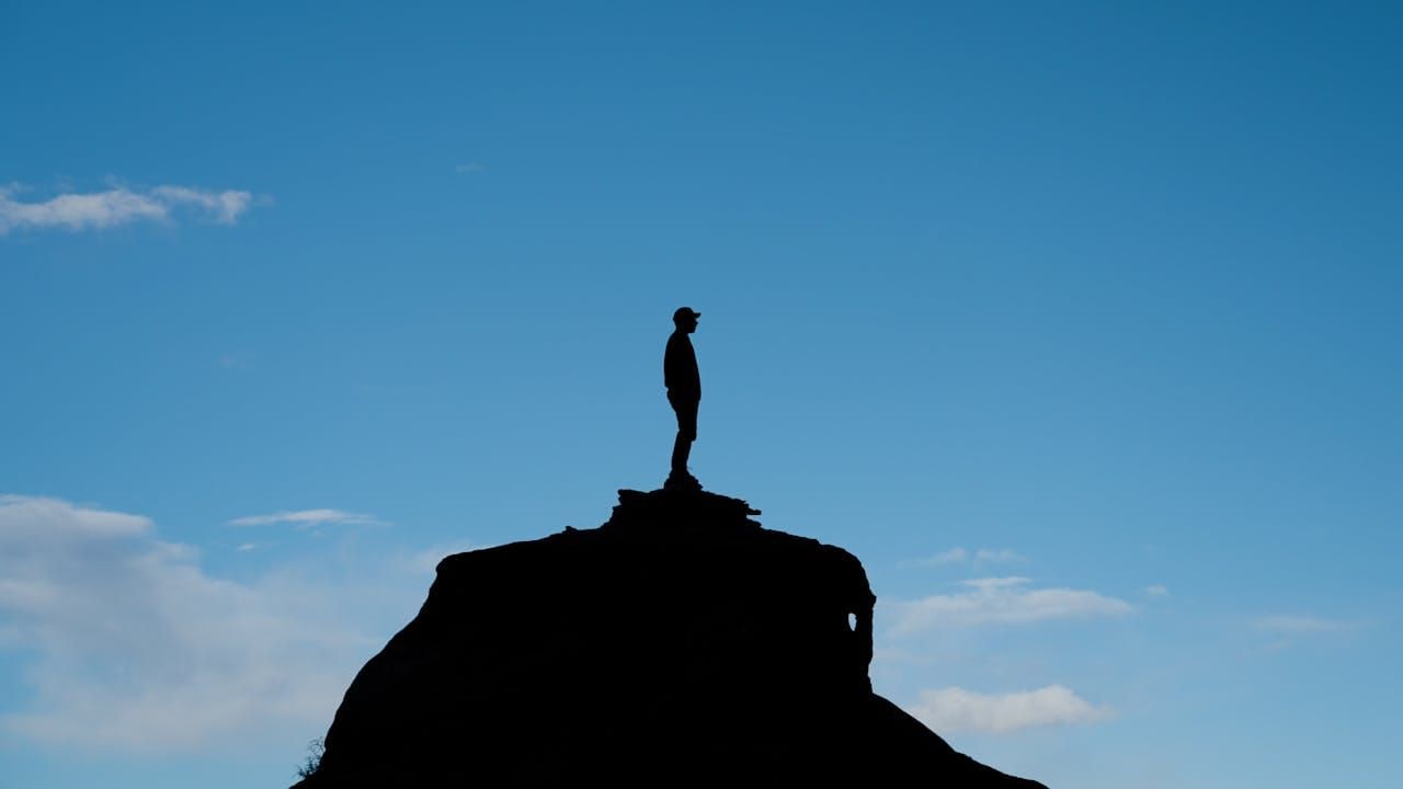 Silhouette of a person standing alone at the top of a hill against a clear blue sky, gazing out into the distance.