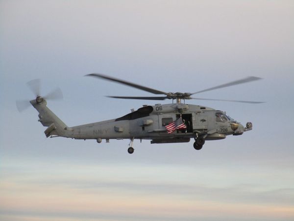 A photo of a helicopter taking off from the deck of a Navy ship with high clouds streaked with purple and pink in the background.