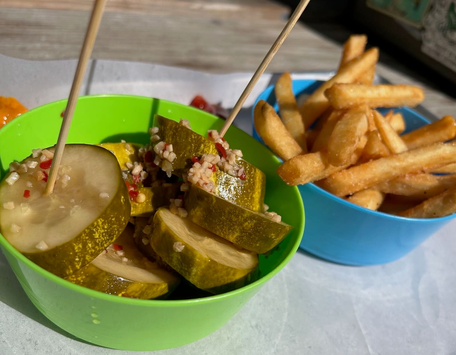 Close-up of garlic pickles with red pepper in green bowl and French fries in blue bowl on picnic table.