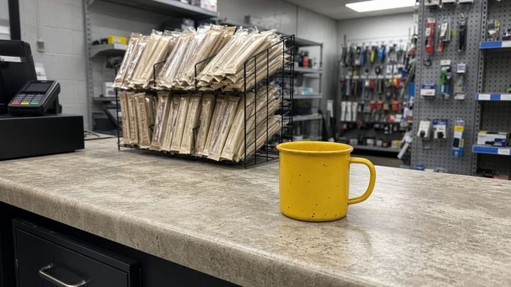 Hardware store checkout counter, a wire snack rack behind the register and a sunflower-yellow enamel mug on the counter, handheld phone photo.