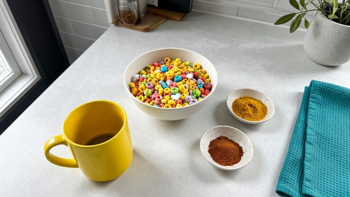 Unlabeled colorful cereal in a white bowl beside turmeric and paprika dishes and a sunflower-yellow mug, handheld iPhone photo in morning window light.